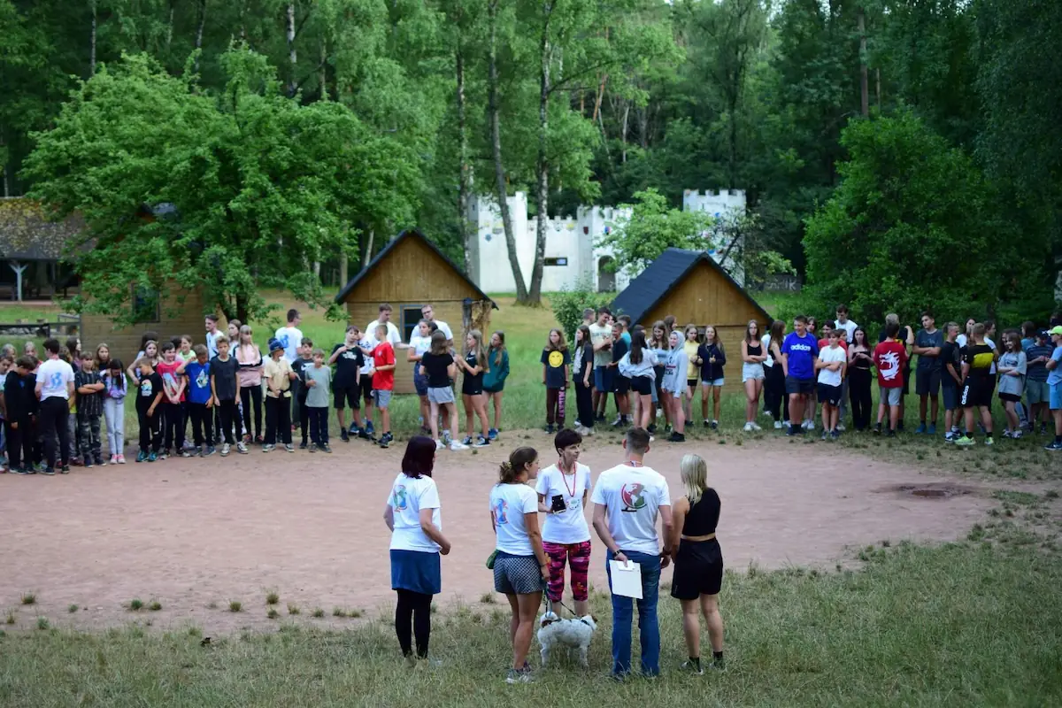 Zaplněné fotbalové hřiště během nástupu tábora. V popředí stojí vedení tábora, 2 vedoucí a dítě.