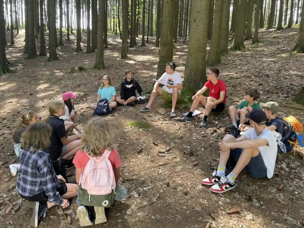 Oddíl dětí se svým vedoucím a praktikantem sedí v lese v kroužku a povídají si.